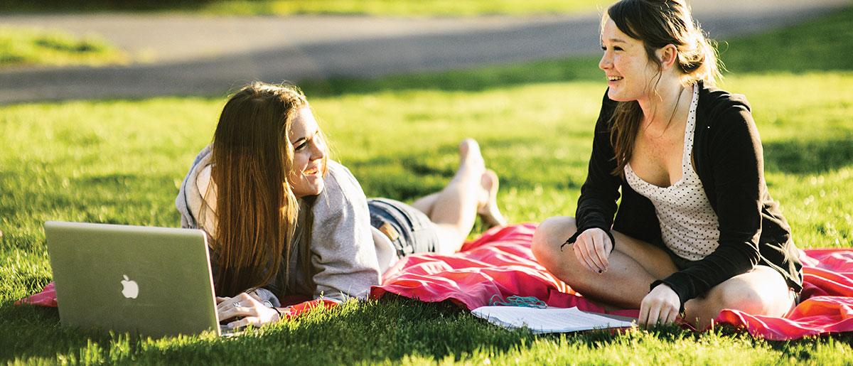 学生s relaxing on the grass, on is laying on a blanket in front of a computer.